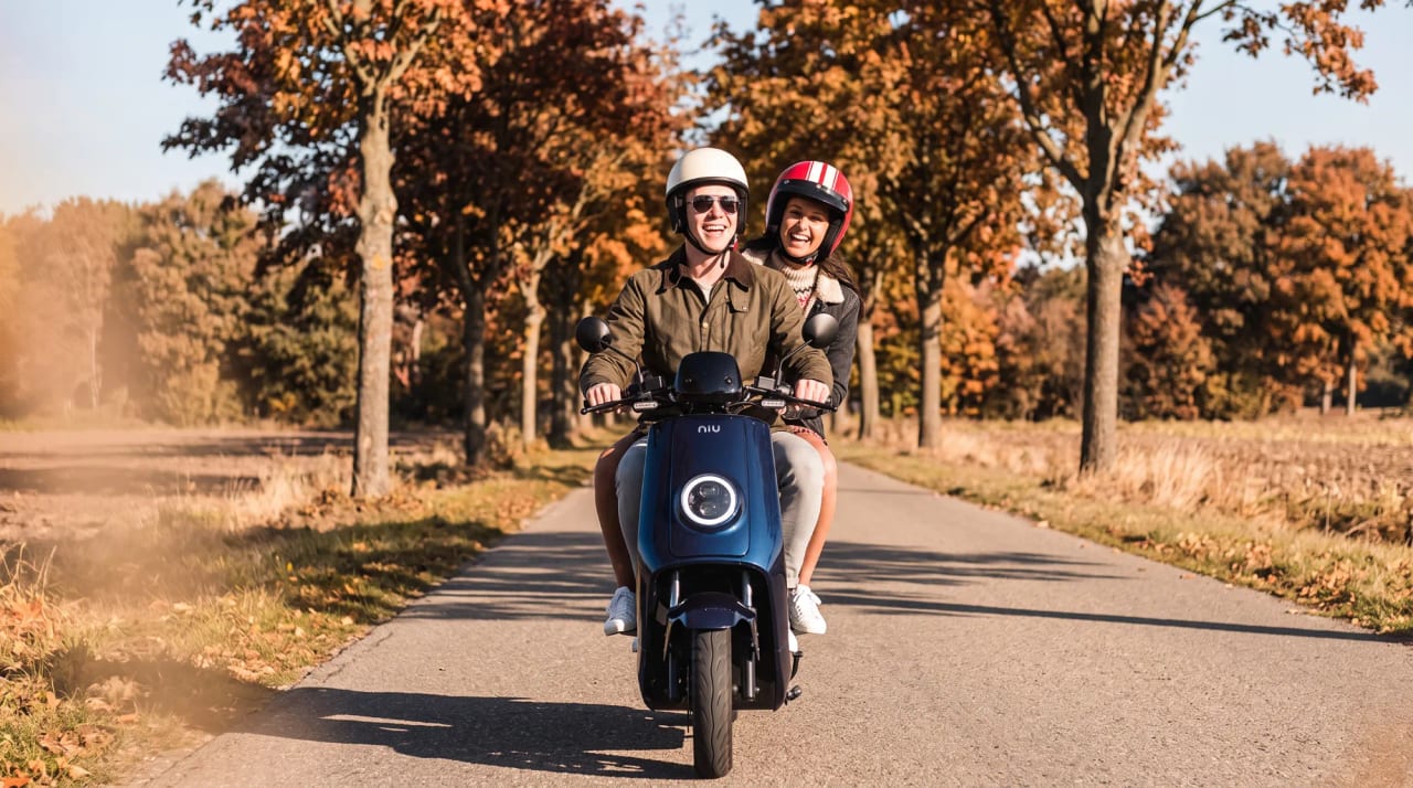 Lachend stel met helmen op een donkerblauwe elektrische scooter op een landweg tussen herfstbomen